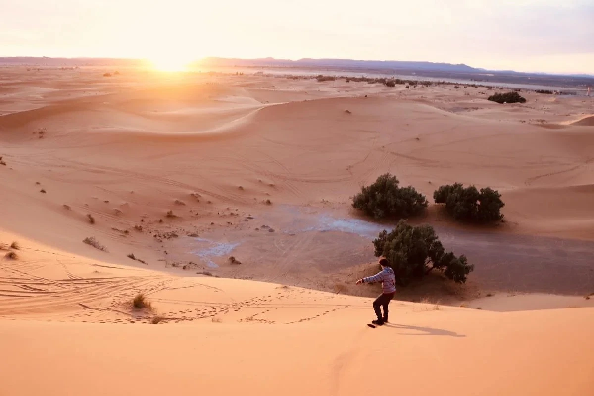 A person is sandboarding down a dune in a vast desert landscape during sunset.