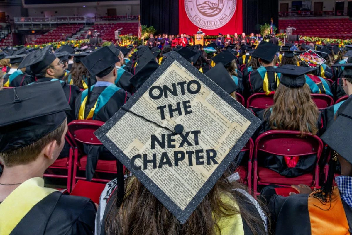 Graduates sit in a commencement ceremony, with one cap decorated with "ONTO THE NEXT CHAPTER."